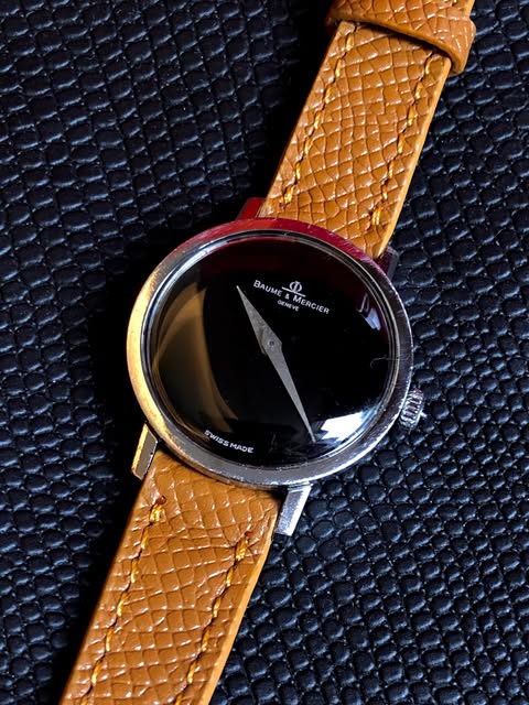 Baume and Mercier watch with an unmarked black dial, triangular hands and a tan textured strap.