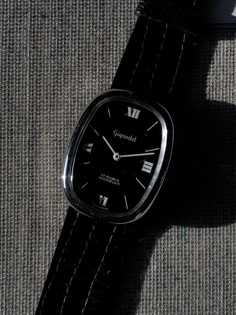 Silver-tone Gigandet watch with a softly rounded elongated case and black dial on woven fabric.