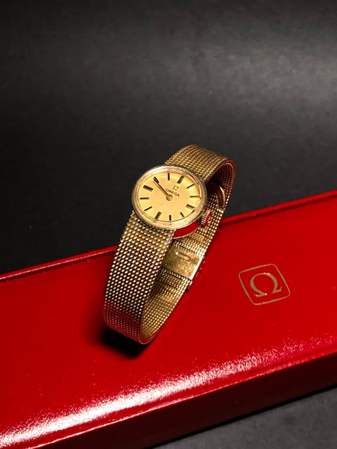 Gold-tone Omega watch with a woven bracelet on a red Omega box.