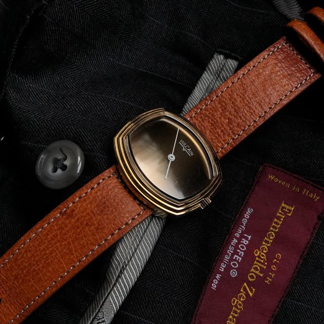 Stepped gold-tone Vulcain watch with a dark dial and brown strap on black wool.